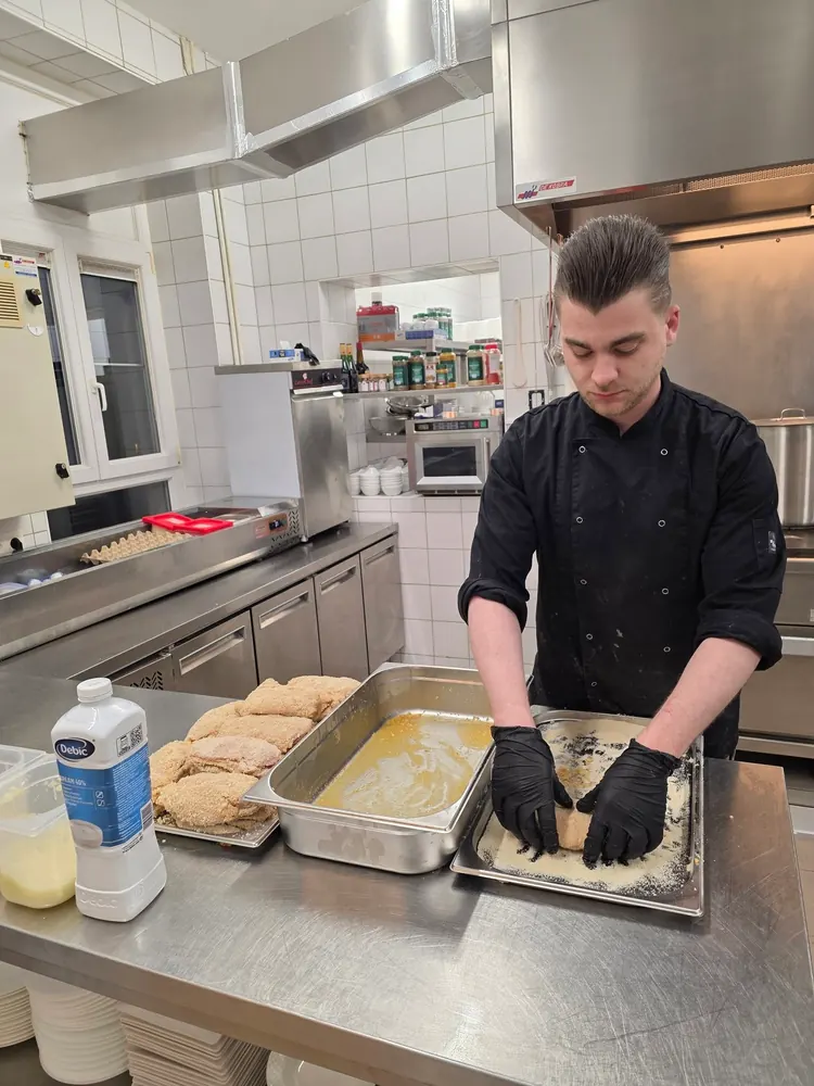 En cuisine, un cuisinier pane à la main des pièces de viande, sur le plan de travail en inox
